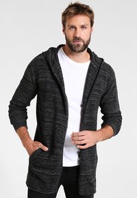 Cárdigan negro texturizado con capucha, mangas largas y fragedo abierto, sobre una camiseta blanca, con dos bolsillos frontales.