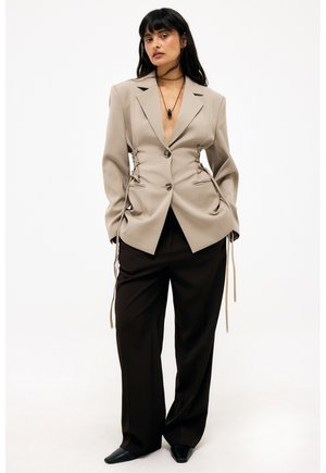 Vrouw in een beige blazer met zijkanten met veters, zwart wijde broek, zwarte puntige schoenen en een zwarte hangersketting tegen een witte achtergrond.