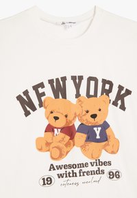 Camiseta blanca de algodón que presenta dos ositos de peluche de dibujos animados vestidos con camisetas rojas y azules con las impresiones "N" y "Y", rodeados de texto y elementos decorativos.