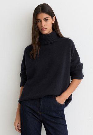 Woman with long dark hair wearing a black turtleneck sweater and dark blue jeans, standing with one hand in pocket against white wall.