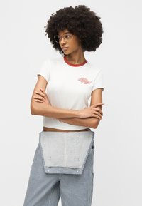 Γυναίκα με σγουρά μαλλιά και γυαλιά, φορώντας λευκή κορυφή crop με κόκκινο περιθώριο και ριγέ παντελόνια με ποδιά, με τα χέρια σταυρωμένα, κοιτάζοντας αριστερά.