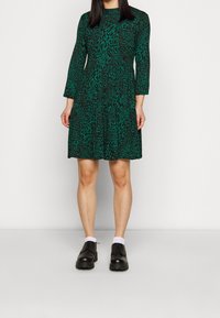 Grön leopardmönstrad klänning med långa ärmar, lagerdesign och rund nacke; bärs med svarta skor och vita strumpor.