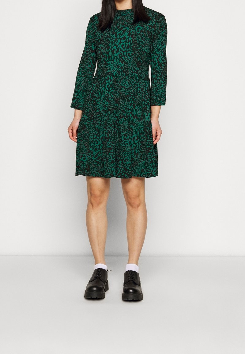 Grön leopardmönstrad klänning med långa ärmar, lagerdesign och rund nacke; bärs med svarta skor och vita strumpor.