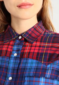 Camicia a quadri rossa e blu con collo classico, chiusura frontale con bottoni e motivi a scacchi contrastanti di larghezze variabili. Tessuto morbido.