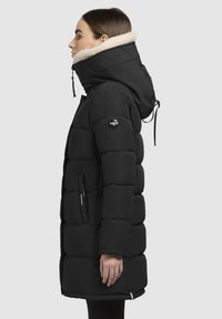 Schwarze gepolsterte Jacke mit hohem Kragen, ausgestattet mit einem cremefarbenen Lammfellbesatz, Reißverschlusstaschen an den Seiten und einem runden Logo-Patch am Ärmel.