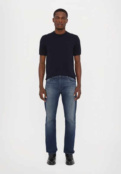 Marineblauw T-shirt met korte mouwen in combinatie met medium-wash straight-leg jeans en zwarte schoenen, staande tegen een witte achtergrond.