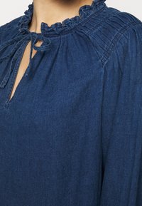 Niebieska bluzka z denimu z falbanką przy dekolcie, wycięciem typu keyhole z przodu oraz marszczeniami na ramionach. Miękka w dotyku, o luźnym kroju.