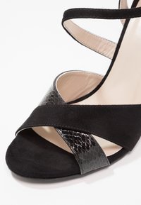 Sandale à talons hauts noire, combinant suède et un matériau embossé noir brillant, avec des lanières croisées et un bout fermé.