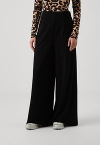 Black wide-leg trousers with front pleats, paired with a leopard print top. The model wears iridescent sneakers, featuring a textured fabric.