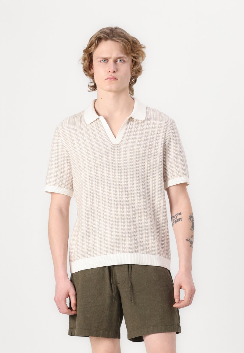 Kortærmet polo i struktureret beige strik, med hvid krave og kant. Båret med olivenfarvede shorts.