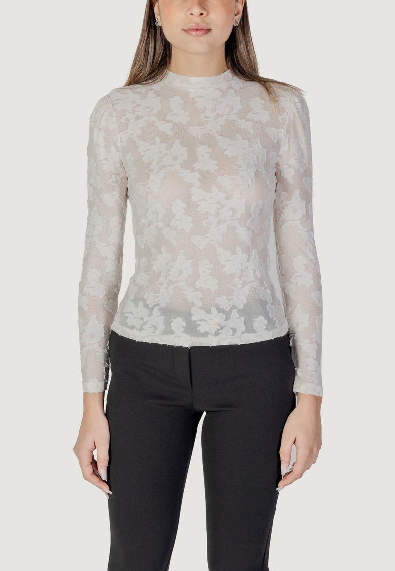 Maglietta bianca a maniche lunghe con sovrapposizione di pizzo floreale, con una silhouette aderente e scollo alto, abbinata a pantaloni neri sartoriali.