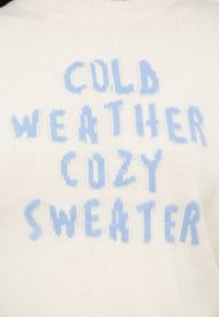 Valge kootud kampsun, millel on sinised plokk tähed, mis loevad „KÜLM ILM SOE KAMPSUN“. Sile tekstuur ja klassikalise ümara kaelusega.