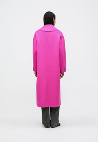 Cilvēks ar tumšiem cirtainiem matiem, ģērbies garā spilgti rozā mētelī, melnās biksēs un melnās kurpēs, stāv uz vienkārša balta fona.