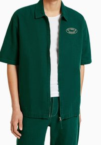 Camisa verde de manga corta con cuello, cierre frontal y parche bordado. Tejido ligero con una textura suave, combinada con pantalones a juego.