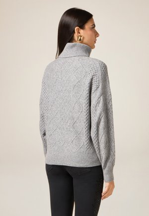 Grau gestrickter Pullover mit hohem Kragen, der ein Rautenmuster und gerippte Bündchen aufweist. Kombiniert mit schwarzer Jeans und präsentiert eine lockere Passform.
