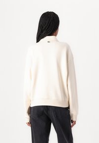 Kremfarget genser med avslappet passform, ribbestrikkede mansjetter og nederkant. Har en liten grønn logo på baksiden av nakken. Kombinert med svarte jeans.