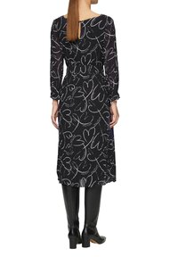 Schwarzes Kleid mit weißem Herzmuster, langen Ärmeln und Rückenschlitz; kombiniert mit kniehohen schwarzen Lederstiefeln.