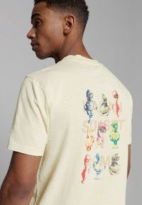 Lichtgeel katoenen t-shirt met een kleurrafte afbeelding van hoofdsculpturen met abstracte patronen op de achterkant. Korte mouwen, ronde hals.