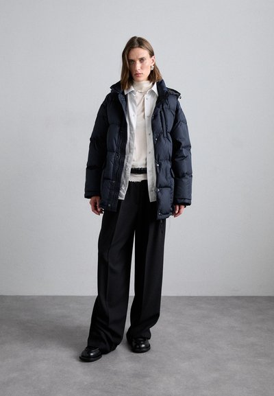 Marineblauwe pufferjack met een afneembare capuchon, gecombineerd met een witte blouse en brede zwarte broeken van gestructureerde stof.
