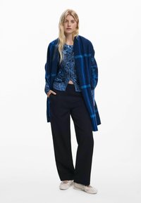Chemise bleue à motifs sous un cardigan ouvert texturé bleu, associée à un pantalon noir ample et des chaussures de couleur claire.