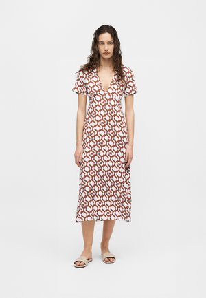 Žena stojaca v midi šatách s krátkym rukávom a výstrihom do tvaru V, hnedej a bielej vzorovanej farby, s béžovými sandálmi na jednoduchom pozadí.