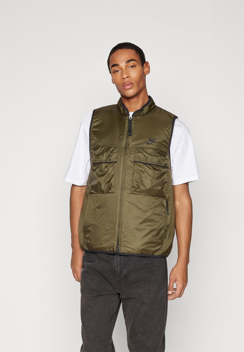 Gilet en nylon vert olive avec une fermeture éclair sur le devant, col montant, deux poches avant et un petit logo. Porté sur un t-shirt blanc et un pantalon noir.