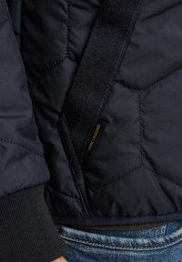 Navy gesteppte Jacke mit gerippten Bündchen, ausgestattet mit einem strukturierten Riemen und einem schwarzen Etikett mit der Aufschrift "Prime Legend." Der Stoff ist glatt und isolierend.
