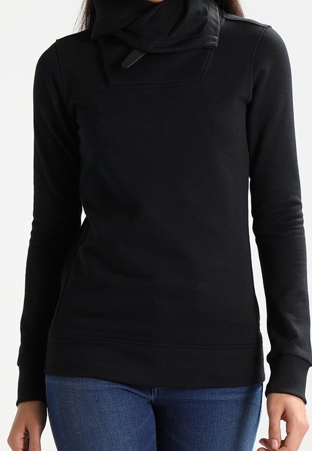 Femme portant un haut noir à manches longues avec un col haut et un détail boucle, associée à un jean bleu, debout devant un fond uni.