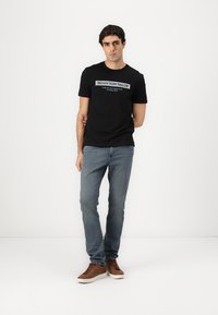 Čierne tričko s krátkym rukávom s logom „DENIM TOM TAILOR“ vpredu, v kombinácii so svetlými modrými džínsami a hnedými teniskami. Jednoduché biele pozadie.