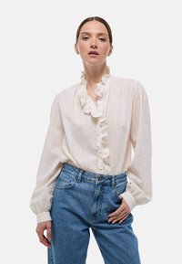 Blouse blanche avec une texture subtile, un col et des poignets volants, et une fermeture à boutons sur le devant, associée à un jean bleu taille haute.