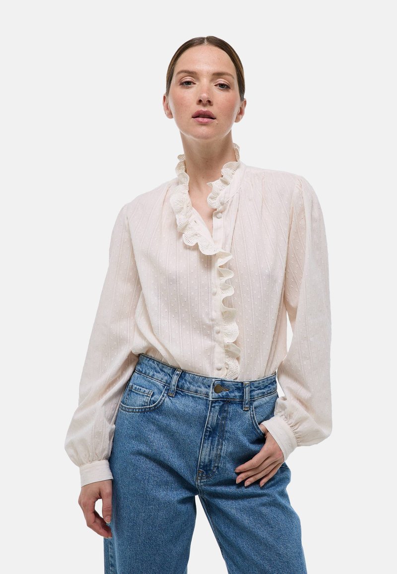 Blouse blanche avec une texture subtile, un col et des poignets volants, et une fermeture à boutons sur le devant, associée à un jean bleu taille haute.