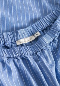 Lichtblauwe stof met witte verticale strepen, met een elastische tailleband en een zichtbaar label met de tekst "STUDIO FEDER" en maat "S."