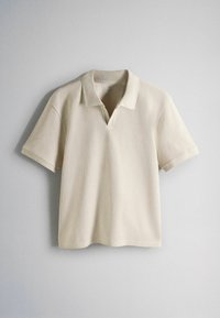 Bézs rövid ujjú kötött galléros póló V-alakú nyakkivágással, sima világosszürke fal előtt lógva.