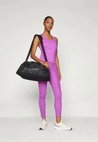 Juodas sportinis krepšys su baltais akcentais, rožinės spalvos marškinėliai be rankovių ir violetiniai šabloniniai leggingai. Modelis, dėvintis baltus sportinius batelius, stovi neutraliame fone.