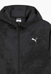 Jachetă neagră ușoară cu un finisaj texturat, fermoar complet pe față și un logo alb Puma pe partea stângă a pieptului. Glugă ajustabilă și interior neted.
