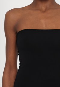 Femme portant un haut noir sans bretelles, montrant les épaules, le haut de la poitrine et les bras sur un fond clair uni.