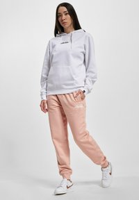 Hvit bomullshoodie med lomme foran og svart logo; rosa joggebukser med elastiske mansjetter og hvit logo; hvite joggesko med brune detaljer.