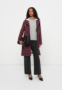 Parka à prova d'água na cor bordeaux com capuz, suéter cinza, calças largas pretas e bolsa preta; modelo a usar saltos altos pretos pontiagudos.