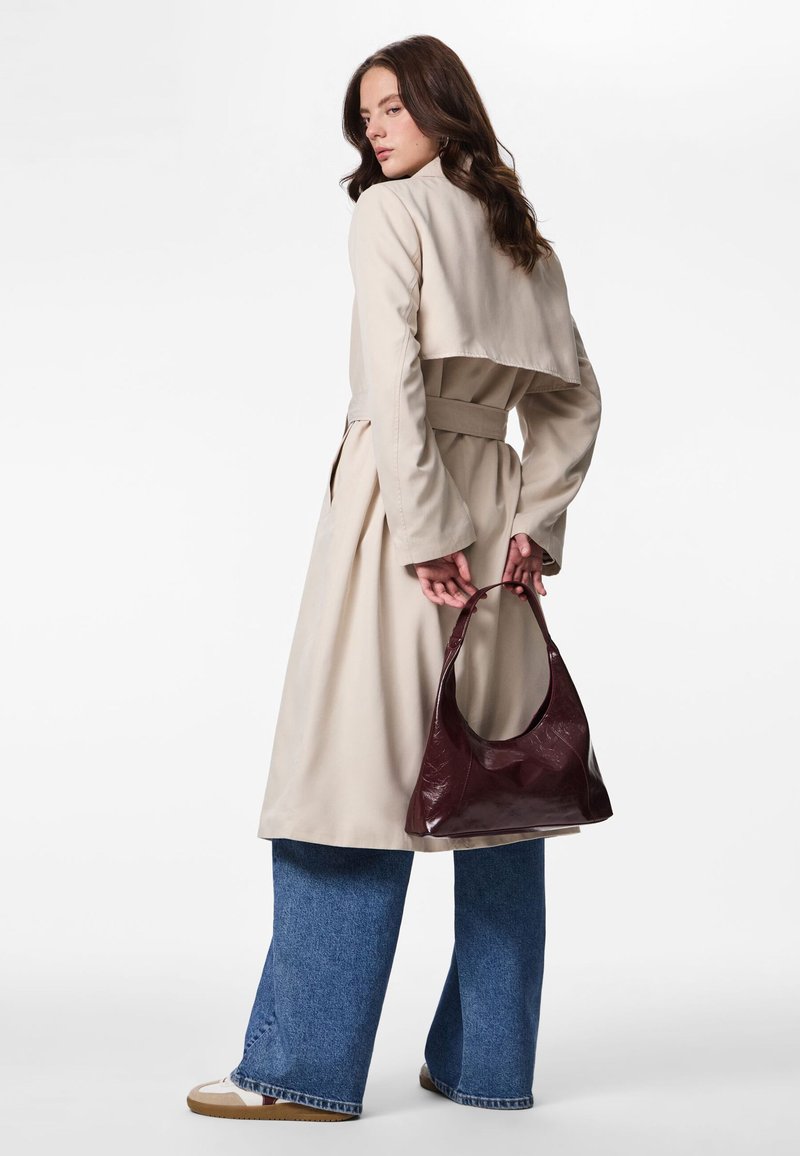 Frau trägt beige Trenchcoat und weite blaue Jeans, hält eine dunkelburgunderfarbene Handtasche hinter dem Rücken, steht vor weißem Hintergrund.