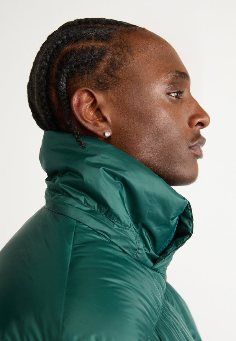 Zielona ocieplana kurtka z wysokim kołnierzem, błyszcząca powierzchnia i widoczne szwy. Zawiera zbliżenie na profil z plecionymi włosami i kolczykami.