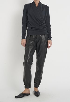 Personne portant un haut cache-cœur à manches longues noir, un pantalon jogger en cuir noir avec des poignets élastiques, et des ballerines noires à bout pointu, debout sur un sol blanc.