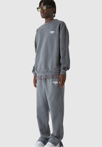 Szürke pulóver és hozzáillő melegítőnadrág puha anyagból, bordás mandzsettákkal és egy kis fehér logóval a mellrészen és a lábon.