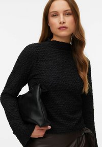 Vrouw met lang bruin haar draagt een zwart textuur top met lange mouwen en houdt een zwarte leren clutch vast tegen een witte achtergrond.