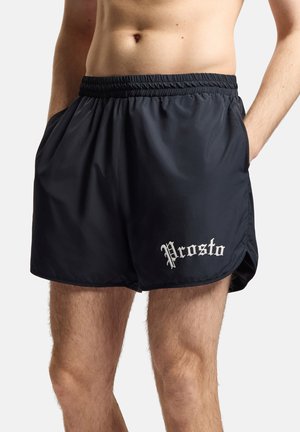 Mężczyzna ubrany w czarne sportowe szorty z elastycznym pasem i białym napisem „Prosto” na lewej nodze, ręce w kieszeniach, na jednolitym tle.