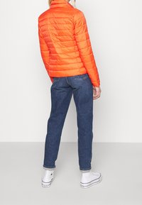 Orange quiltet puffjakke med opretstående krave, kombineret med blå jeans med lige ben og hvide høje sneakers. Enkel, funktionel design.