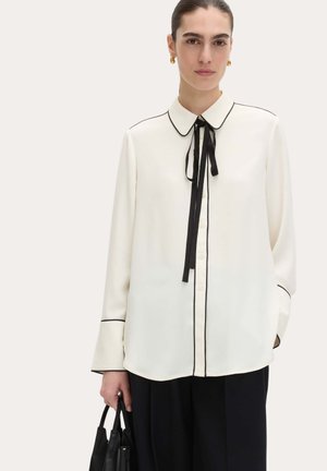 Mujer con una blusa blanca con ribete negro y un lazo negro, pantalones negros, pendientes de aro dorados, sosteniendo un bolso negro.