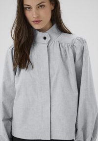 Kvinde med langt brunt hår iført en lys grå knap-op højhalset bluse med pufferede skuldre mod en ensfarvet baggrund.