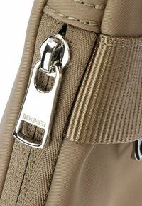 Beige nylon bag featuring a silver zipper pull with engraving, textured fabric, and a visible seam along the edge.