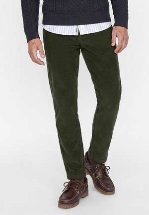Homme portant un pantalon en velours côtelé vert foncé, des chaussures en cuir marron foncé, un pull bleu marine et une chemise blanche rayée, debout sur un fond blanc.