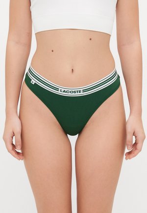 Gröna bomullstrosor med vitrandig resårmidja som har "LACOSTE"-logotypen. Slät textur och åtsittande design.